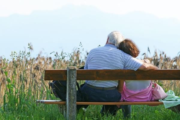 Zwei von hinten sichtbare Senioren sitzen auf einer Bank und umarmen sich